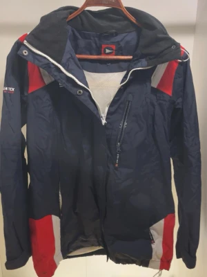 Marinblå vindjacka med röda detaljer - Snygg marinblå vindjacka från Aqua Tex med röda och vita detaljer. Jackan har huva, dragkedja framtill, bröstficka med dragkedja och meshfoder. Perfekt för blåsiga dagar och sportiga outfits.