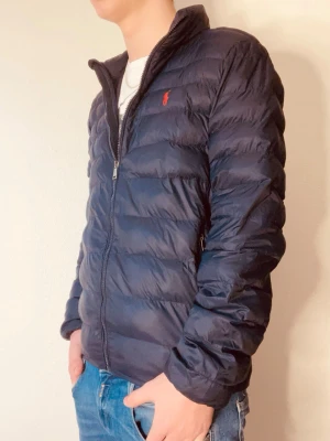 Mörkblå dunjacka från Ralph Lauren - Snygg mörkblå dunjacka från Ralph Lauren med diskret röd logga på bröstet. Jackan har dragkedja framtill, två sidofickor med dragkedja och en klassisk ståkrage. Perfekt för kalla vinterdagar och riktigt lätt att bära.