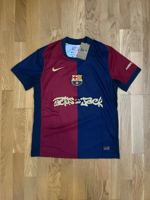 Barcelona x Nike Travis Scott matchtröja - Säljer en officiell FC Barcelona matchtröja i samarbete med Travis Scott och Nike. Tröjan är blå och vinröd med klubbmärke, Nike-logga och Travis Scott-grafik på bröstet. Tillverkad i lätt och ventilerande material, perfekt för fotboll eller streetwear.