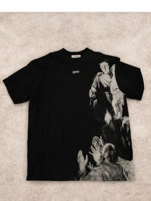 Svart Off-White t-shirt med print - Svart t-shirt från Off-White med vit text 'OFF' på bröstet och ett stort konstnärligt print i vitt på framsidan. Baksidan har Off-White-loggan i vitt. Klassisk rund hals och korta ärmar. Materialet är mjuk bomull som känns skön mot huden. Finns med prislapp. Äkta 