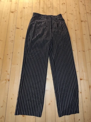 Svarta kostymbyxor med vita ränder randig - Snygga svarta kostymbyxor med vita vertikala ränder och hög midja. Byxorna har raka ben och avslappnad passform, perfekta för en trendig och stilren look. Passar dig som gillar klassiska men ändå moderna detaljer.