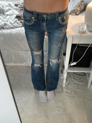 Blåa bootcut jeans från Miss Me - Säljer ett par blåa bootcut jeans från Miss Me med slitningar och hål på benen. Jeansen har dekorativa bakfickor med broderier och nitar samt detaljerade sömmar. Klassisk femficksmodell och låg midja. Perfekta för en trendig och avslappnad stil. 36cm raktöver midjan och passar bra på mig som har xs/s och är 158. Dom är avklippta längst ner.