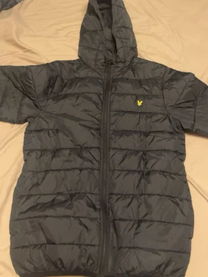 Svart dunjacka med huva från Lyle & Scott - Säljer en svart dunjacka från Lyle & Scott med huva och dragkedja framtill. Jackan har quiltad design och den klassiska gula loggan på bröstet. Perfekt för kalla vinterdagar och riktigt skön att bära. Materialet är syntet med dunfyllning.