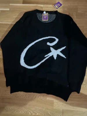 Svart stickad tröja by Corteiz  - Cool svart stickad tröja från Corteiz med stor vit C-logga framtill. Tröjan har rund halsringning och ribbade muddar vid ärmslut och nederkant. Perfekt för dig som gillar streetwear och vill sticka ut lite extra.