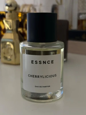 ESSNCE Cherrylicious Eau de Parfum - Minimalistisk parfymflaska från ESSNCE med doften Cherrylicious. Flaskan är cylindrisk i genomskinligt glas med svart matt kork och enkel vit etikett med svart text. Innehållet är ljust gult och flaskan har en modern, stilren design.