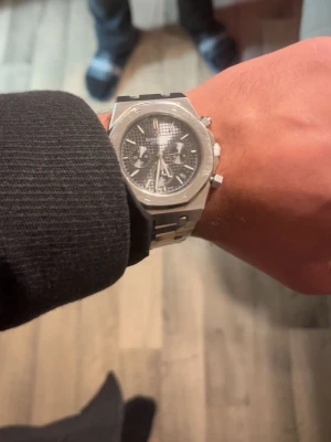Audemars Piguet Royal Oak klocka - Snygg Audemars Piguet Royal Oak klocka i silver med svart urtavla och klassiskt rutmönster. Klockan har kronograf, datumvisning och robust länkarmband i metall. Den ikoniska åttakantiga boetten ger en riktigt exklusiv känsla.
