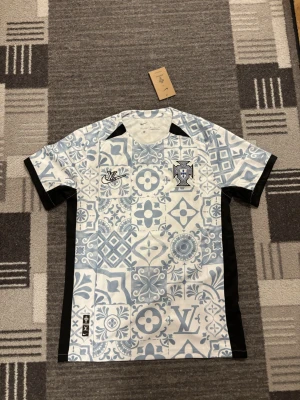 LV x nike jersey - Snygg Portugal matchtröja från Nike i blått och vitt med unikt kakelinspirerat mönster. Tröjan har svarta sidopaneler, rund hals och broderad Portugal-logga på bröstet. Fick den som present och Jag har inte haft på mig den för att den är för stor.