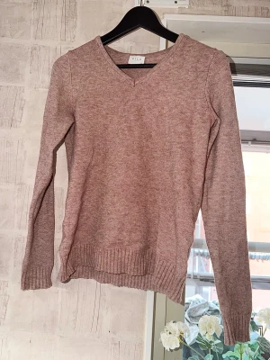 Rosa stickad tröja från Vila - Mysig stickad tröja från Vila i en mjuk, ljusrosa nyans. Tröjan har V-ringning, långa ärmar och ribbade muddar vid ärmslut och nederkant. 
