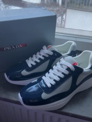 Blå vita prada skor - Snygga Prada skor  i blankt mörkblått och vitt med vita meshpaneler och vita skosnören. Klassisk låg modell med tjock vit sula och röda Prada-detaljer på plösen och sulan. Perfekta för dig som vill ha en exklusiv och trendig look.