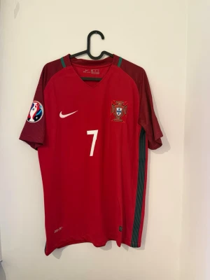 Portugal matchtröja Ronaldo #7 Nike - Snygg röd Portugal matchtröja från Nike med nummer 7 och Ronaldo på ryggen. Tröjan har officiellt landslagsemblem, Nike-logga och EM-märke på ärmen. Tillverkad i lätt och ventilerande material, perfekt för fotboll eller som supporterplagg.