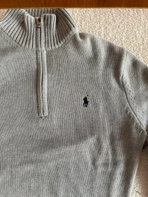 Grå stickad tröja från Ralph Lauren - Snygg grå stickad tröja från Ralph Lauren med halv dragkedja och hög krage. Tröjan har ribbade muddar vid ärmslut och nederkant samt den klassiska broderade loggan på bröstet. Perfekt för lager på lager och stilrena outfits.