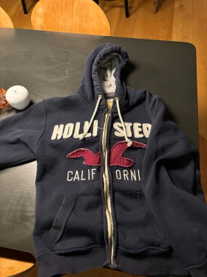 Marinblå Hollister hoodie med dragkedja - Passformen är mer som en M/S än L. Mysig marinblå hoodie från Hollister med dragkedja, broderad logga och text i vitt samt röda detaljer på bröstet. Utrustad med huva och vita dragsnören, samt två fickor framtill. Insidan är fodrad i mjukt material för extra komfort. 