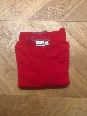 Röd stickad tröja från Calvin Klein - Röd stickad tröja från Calvin Klein med V-ringning och diskret logga på bröstet. Tröjan har gråa detaljer på ärmarna och ribbstickade muddar vid ärmslut och nederkant. Perfekt för dig som gillar klassisk och stilren design.