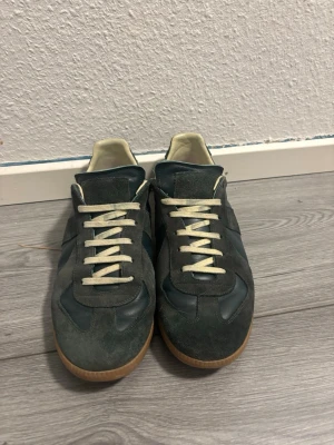 Gröna Maison Margiela Replica sneakers - Snygga gröna Maison Margiela Replica sneakers med ovandel i mocka och läder, vita snören och beige innersula. Klassisk gummisula och låg profil som ger en clean look. Perfekta för dig som gillar stilrena och tidlösa sneakers.
