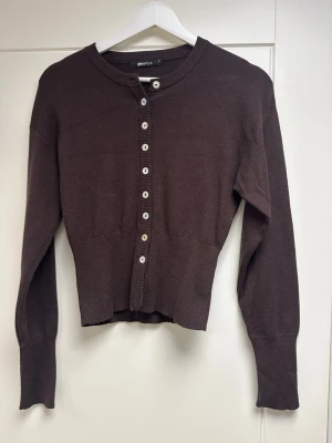 Brun cropped kofta från Gina Tricot S  - Mörkbrun cropped kofta från Gina Tricot med långa ärmar och ribbade muddar. Koftan har rund halsringning och ljusa knappar framtill. .