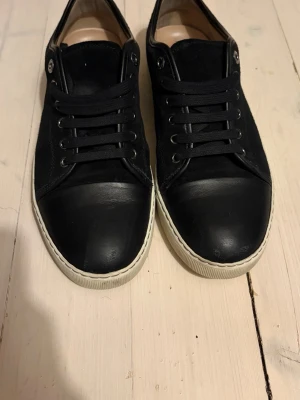 Svarta Lanvin sneakers i mocka och läder - Snygga svarta sneakers från Lanvin med ovandel i mocka och detaljer i läder. Klassisk låg modell med svarta skosnören och metallöljetter. Perfekta för dig som gillar stilrena och exklusiva skor.