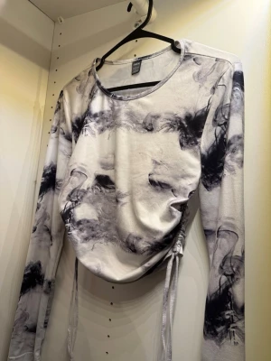 Långärmad topp med svartvitt mönster - Trendig långärmad topp med abstrakt svartvitt mönster och snörning i sidan för en snygg, rynkad effekt. Mjuk och stretchig känsla, perfekt till jeans eller kjol. Rund halsringning och croppad passform ger en modern vibe.