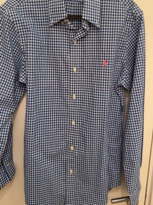 Blårutig skjorta från Ralph Lauren - Klassisk blårutig skjorta från Ralph Lauren med lång ärm och knappar framtill. Skjortan har en liten rosa logga broderad på bröstet och är tillverkad i mjuk bomull. Perfekt för en stilren och avslappnad look.