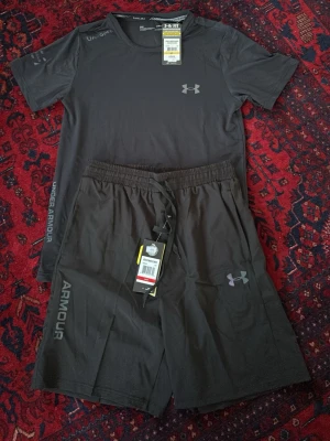 Svart träningsset från Under Armour - Säljer ett svart träningsset från Under Armour med t-shirt och shorts. Båda plaggen har logga på bröst och ben samt tryckta detaljer. Materialet är lätt och snabbtorkande, perfekt för gym eller löpning. Elastisk midja med dragsko på shortsen.
