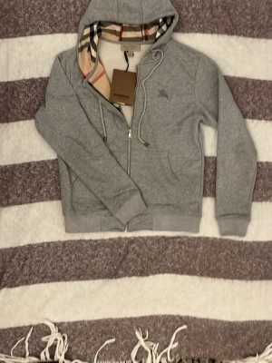 Grå zip hoodie från Burberry - Snygg grå zip hoodie från Burberry med klassiskt rutigt foder i huvan och dragkedja framtill. Hoodien har dragsko i huvan, ribbade muddar och två fickor fram. Perfekt för en chill och stilren look. Materialet är mjukt och bekvämt, troligen bomullsblandning.