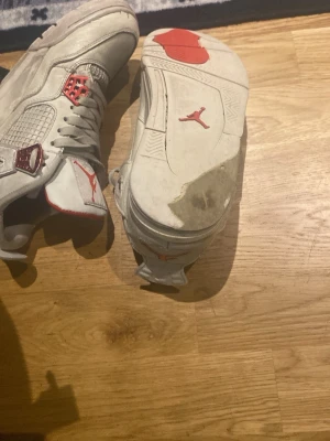 Vita Air Jordan 4 med röda detaljer - Säljer ett par klassiska Air Jordan 4 sneakers i vitt med röda detaljer och meshpaneler. Skorna har högt skaft, ikonisk Jumpman-logga på plös och sula samt röda accenter på snörningen och insidan. Perfekta för dig som gillar streetwear och basketstil.