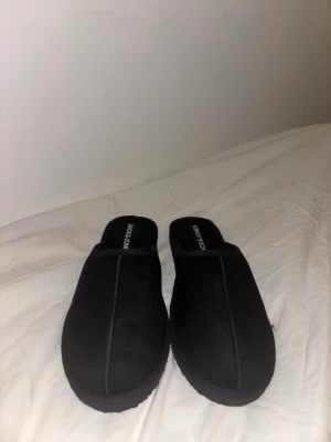 Svarta tofflor från Jack & Jones - Mysiga svarta tofflor från Jack & Jones i klassisk slip-in modell. Tillverkade i mjukt textilmaterial med diskreta sömmar längs ovansidan och bekant logga på innersulan. Perfekta för att chilla hemma och hålla fötterna varma.