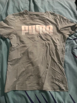 Grå PUMA t-shirt med tryck - Snygg grå t-shirt från PUMA med stort vitt logotyptryck på bröstet. Klassisk passform med rund halsringning och korta ärmar. Perfekt för en sportig och avslappnad stil.