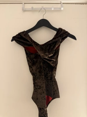 Brun sammetsbody med omlottkrage - Säljer en snygg brun body i sammetsmaterial med omlottkrage och djup urringning. Bodyn har en unik draperad design och insida i röd nyans. Perfekt för dig som vill ha något extra till din outfit.