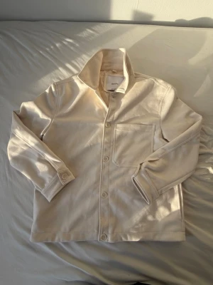 Beige overshirt från Bershka - Stilren beige overshirt från Bershka med klassisk krage, knappstängning framtill och en bröstficka. Tillverkad i ett mjukt tyg som ger en avslappnad look. Perfekt att bära över en t-shirt för en enkel men snygg outfit. Aldrig använt.