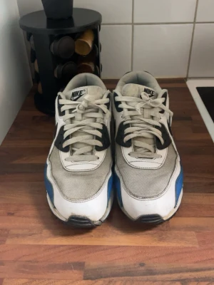Nike Air Max 90 vita/blå sneakers - Nike Air Max 90 sneakers i vitt, grått, svart och blått. Klassisk siluett med mesh och läderdetaljer, synlig Air Max-dämpning i sulan och ikonisk swoosh på sidan. Sköna och tidlösa sneakers som passar till det mesta. Vid snabb köp går priset att sänkas