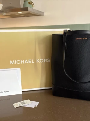 Svart handväska från Michael Kors - Äkta Stilren svart handväska från Michael Kors i slätt och strukturerat läder. Väskan har guldfärgade detaljer och logga framtill samt dubbla handtag. Perfekt för dig som gillar klassisk och tidlös design med lyxig känsla.