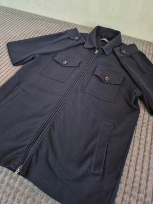 Dressman overshirt med dragkedja - En snygg ät mörkblå overshirt med korta ärmar, dragkedja framtill och två bröstfickor med lock och knapp. Skjortkrage och axelklaffar med knappdetaljer ger en cool militärinspirerad look. Perfekt att bära öppen eller stängd över en t-shirt.