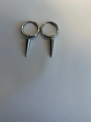 Silvriga hoops med konformad hängare - Säljer ett par silvriga örhängen i form av hoops med konformade hängen. Örhängena har en minimalistisk och edgy stil, perfekta för att ge din look en cool touch. Materialet ser ut att vara metall med blank finish.
