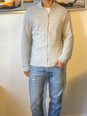 Ljusgrå ziptröja med hög krage - Snygg ljusgrå ziptröja med hög krage och ribbade muddar. Tröjan har en minimalistisk look och är tillverkad i ett mjukt, tunt material som passar perfekt till jeans. Dragkedja framtill och diskret passform.