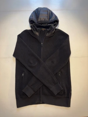 Svart stickad cardigan jacka - Snygg svart stickad cardigan jacka med dragkedja framtill och två fickor med dragkedja. Huva i glansigt material ger en cool kontrast till resten av tröjan. Perfekt för dig som gillar en sportig och avslappnad stil. Storlek M. Skick 9/10.