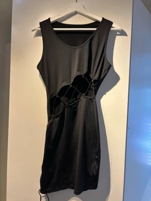 Svart kort klänning med snörning och cut out  - Säljer en svart, ärmlös kort klänning med unik snörningsdetalj över midjan. Klänningen har en tight passform och är tillverkad i ett stretchigt syntetmaterial. Perfekt för dig som vill sticka ut med en edgy och trendig look. Aldrig använd 