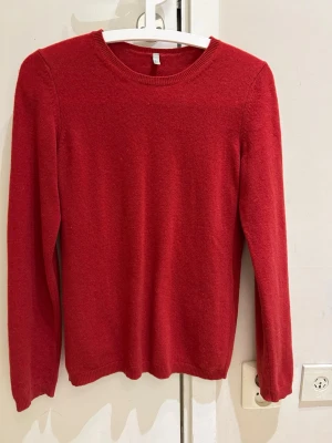 Röd Kashmir stickad tröja från Benetton - Röd stickad tröja från Benetton med rund halsringning och långa ärmar. Tröjan har en klassisk, enkel design och ribbstickade muddar vid hals, ärmslut och nederkant. Perfekt för lager-på-lager under kyliga dagar.