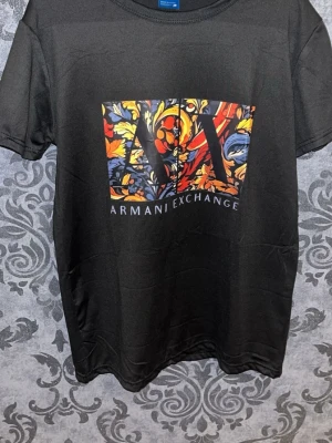 Svart t-shirt från Armani Exchange - Svart t-shirt från Armani Exchange med stort färgglatt tryck framtill i orange, gul, blå och röd med blommigt mönster och AX-logga. Klassisk rund hals och korta ärmar. Perfekt för dig som vill sticka ut med en snygg statement-tee.