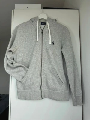 Grå zip hoodie från Polo Ralph Lauren - Snygg ljusgrå hoodie med dragkedja från Polo Ralph Lauren. Klassisk design med huva och vita dragsnören, två fickor framtill och den ikoniska mörkblå loggan broderad på bröstet. Perfekt för en chill och avslappnad stil.