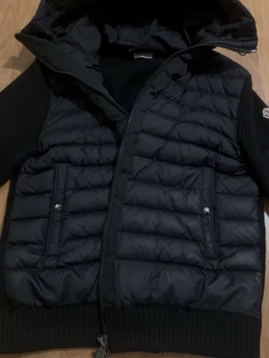 Svart dunjacka från Moncler - Snygg svart dunjacka från Moncler med stickade ärmar och huva. Jackan har två framfickor med tryckknappar och en extra ficka med dragkedja på ärmen. Klassisk Moncler-logga på vänster ärm. Perfekt för kalla vinterdagar.