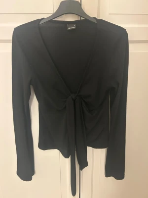 Svart omlottopp från Gina Tricot - Säljer en svart omlottopp från Gina Tricot i storlek S. Toppen har långa ärmar, djup v-ringning och knyt framtill. Perfekt att styla med höga jeans eller kjol för en trendig look.