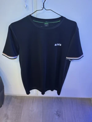 Svart BOSS t-shirt med logga - Svart t-shirt från BOSS med vit logga på bröstet. T-shirten har rund halsringning och korta ärmar med snygga ränder i vitt och beige vid ärmsluten. Perfekt för en stilren och modern look.