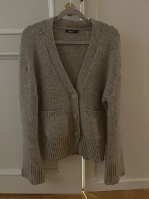 Beige kofta från Gina Tricot - Mysig beige kofta från Gina Tricot med djup V-ringning, tre knappar framtill och två stora fickor. Koftan är stickad med breda ärmar och superfin! Säljer då den inte kommer till användning!