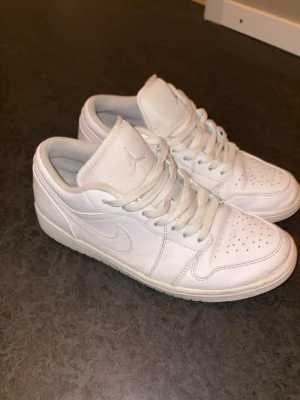 Vita Air Jordan 1 Low sneakers - Snygga helvita Air Jordan 1 Low sneakers i läder med klassisk siluett, perforerad tå och ikoniska Jumpman-loggan på plösen. Skorna har vita snören och diskret Nike Swoosh på sidorna. Perfekta för en clean och stilren look.
