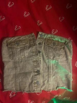 Beige denim tubtopp med knappar - Säljer en beige tubtopp i denim med knappar framtill och två bröstfickor med lock. Toppen har råa kanter nedtill och smockad baksida för skön passform. Perfekt för dig som gillar en edgy och unik stil.