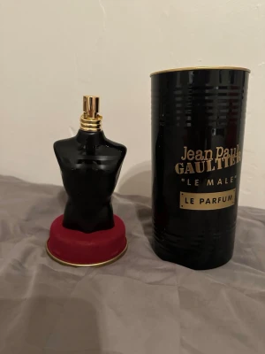 Jean Paul Gaultier Le Male Le Parfum - Snygg parfymflaska formad som en svart manlig torso med gulddetaljer från Jean Paul Gaultier. Kommer i en elegant svart metalltub med guldfärgad text och lock. Flaskan står på en rund röd sammetsliknande bas. Perfekt för dig som gillar lyxiga och unika dofter.