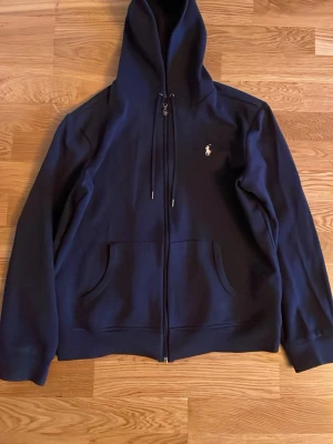 Marinblå Polo Ralph Lauren zip hoodie - Snygg marinblå hoodie från Polo Ralph Lauren med klassisk vit broderad logga på bröstet. Tröjan har huva med snörning, dragkedja framtill och två stora fickor. Tillverkad i mjuk bomull, perfekt för chill dagar eller när du vill ha en clean look.