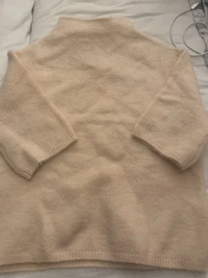 Beige stickad tröja med halvpolo - Mysig beige stickad tröja med halvpolo och trekvartsärmar. Tröjan har en rak passform och är tillverkad i ett mjukt, fluffigt material som känns skönt mot huden. Perfekt för lager på lager och enkel att matcha med olika outfits.