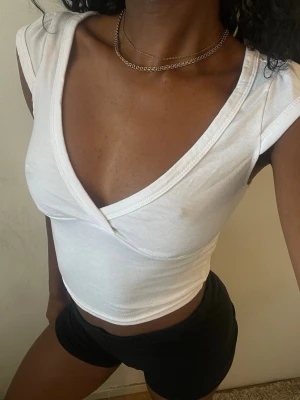 Vit croppad omlottopp - Snygg vit croppad topp med omlottdesign och djup v-ringning. Toppen är ärmlös och har en tight passform som framhäver figuren. Perfekt att styla med jeans eller kjol för en trendig look.