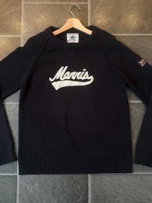 Svart stickad tröja Morris - Svart stickad tröja från Morris med vit broderad logga på bröstet och en liten brittisk flagga på ena ärmen. Tröjan har rund halsringning, långa ärmar och ribbade muddar. Perfekt för en stilren och sportig look. Ska tydligen vara L men passar M lika bra, kolla bild med mått. Äkta!